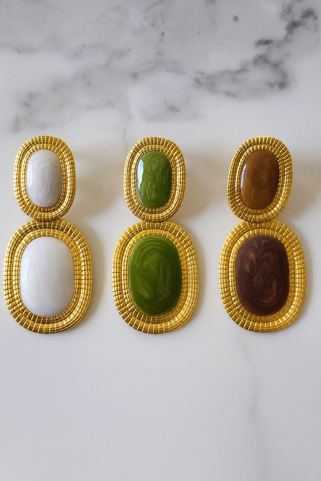 Trois paires de boucles d’oreilles Likasi en acier doré et résine marbrée, présentées côte à côte en blanc, vert et marron.