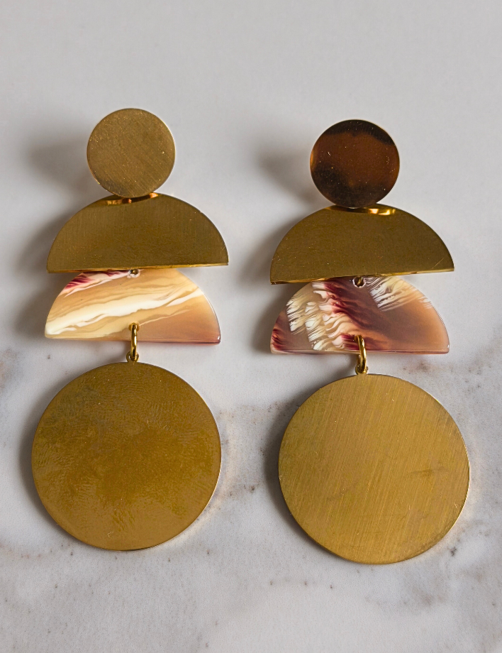 Boucles d’oreilles Coumba en acier doré avec pièce centrale en résine imitation pierre naturelle, forme géométrique longue de 7 cm, posées sur fond marbré clair – Bijoux MOUN