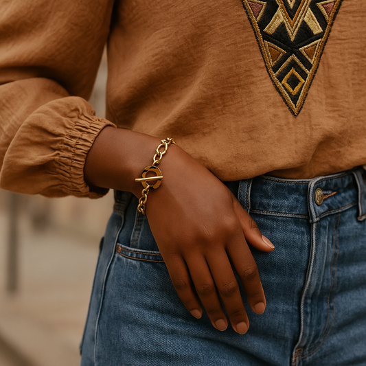 Bracelet Ndomo porté par une femme noire, main posée sur un jean, bracelet en acier doré avec fermoir écaille, style afrochic éditorial.