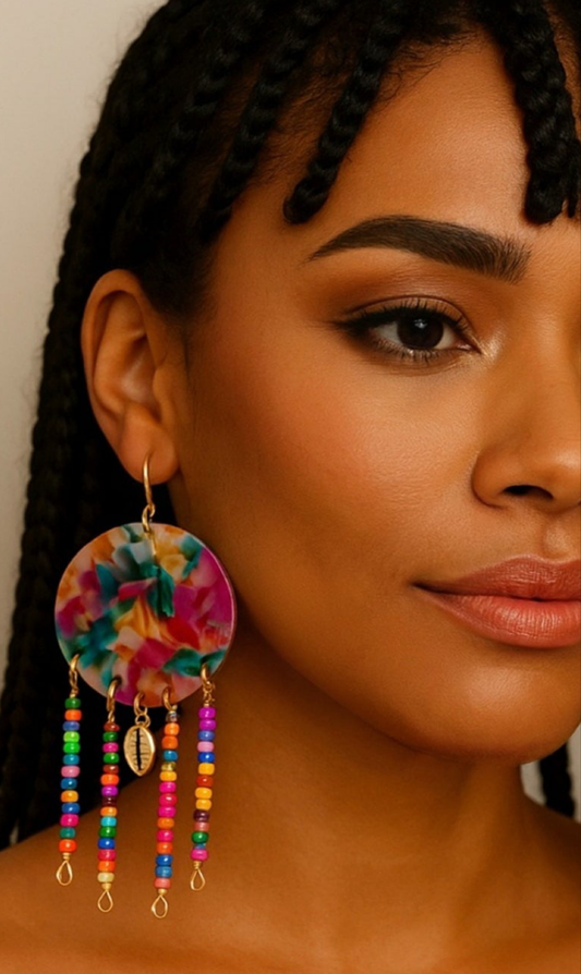Boucles d’oreilles Bahari en résine colorée multicolore, avec pendentifs perles et cauri doré, portées par une femme à la peau mate et aux tresses, style afro-chic.

